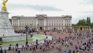 Aerial view of runners participating in the Royal Parks Half Marathon passing in front of Buckingham Palace, with spectators lining the route and the Victoria Memorial visible.