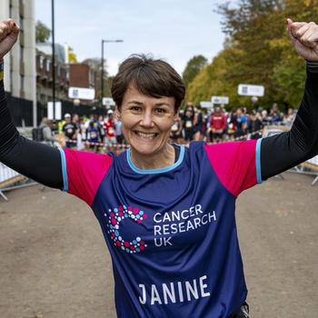 Person celebrating at the start line of the Royal Parks Half, wearing a Cancer Research UK t-shirt with the name "Janine" on it, with spectators and barriers in the background.