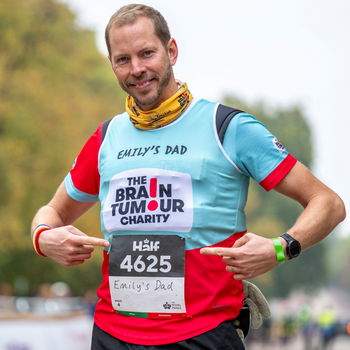 A person is smiling and pointing at a race bib labeled "Emily's Dad." The shirt displays "The Brain Tumour Charity." 